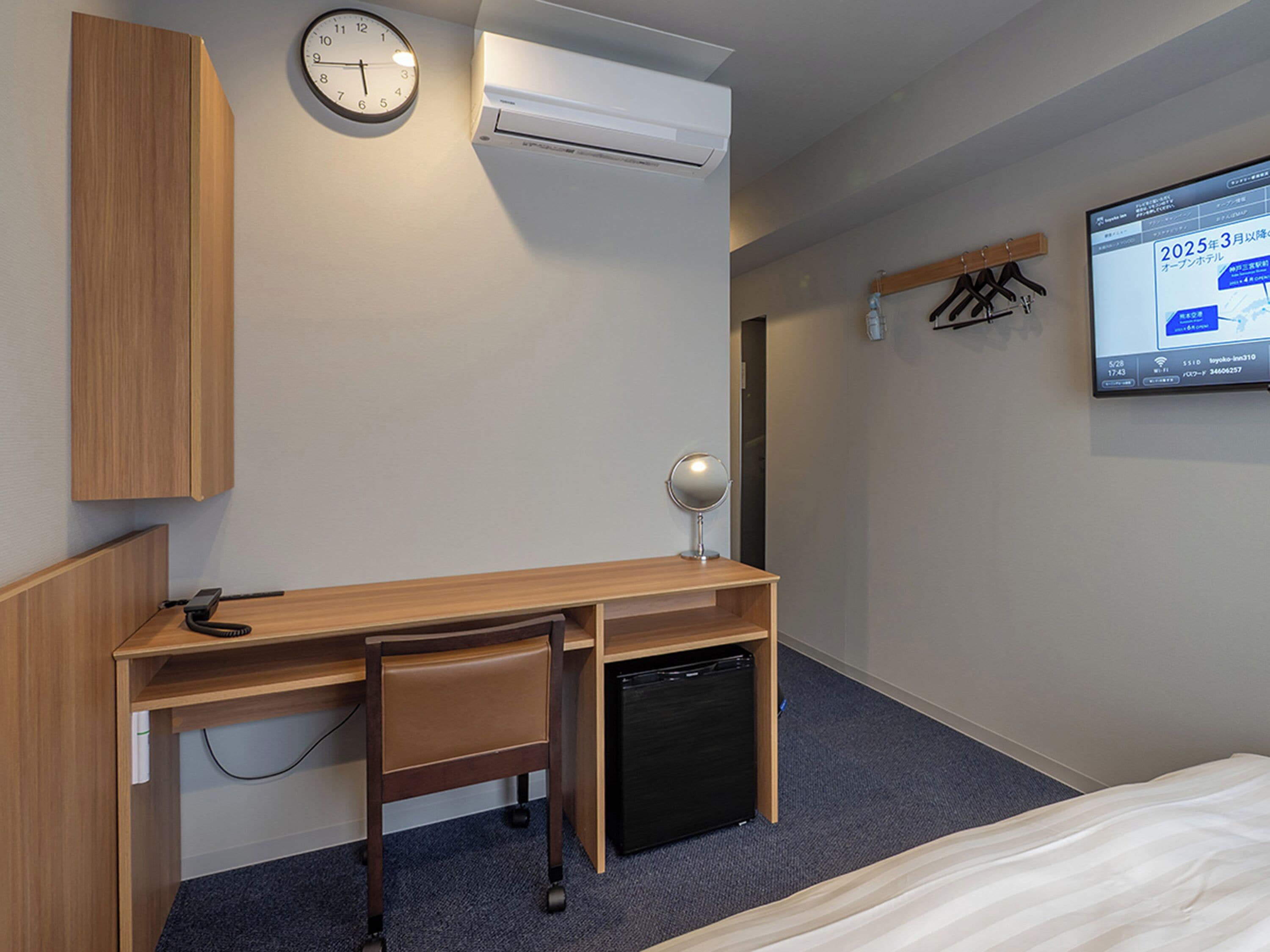 Una camera d'albergo con una scrivania in legno, uno specchio, una sveglia, un condizionatore d'aria, una TV e un letto.