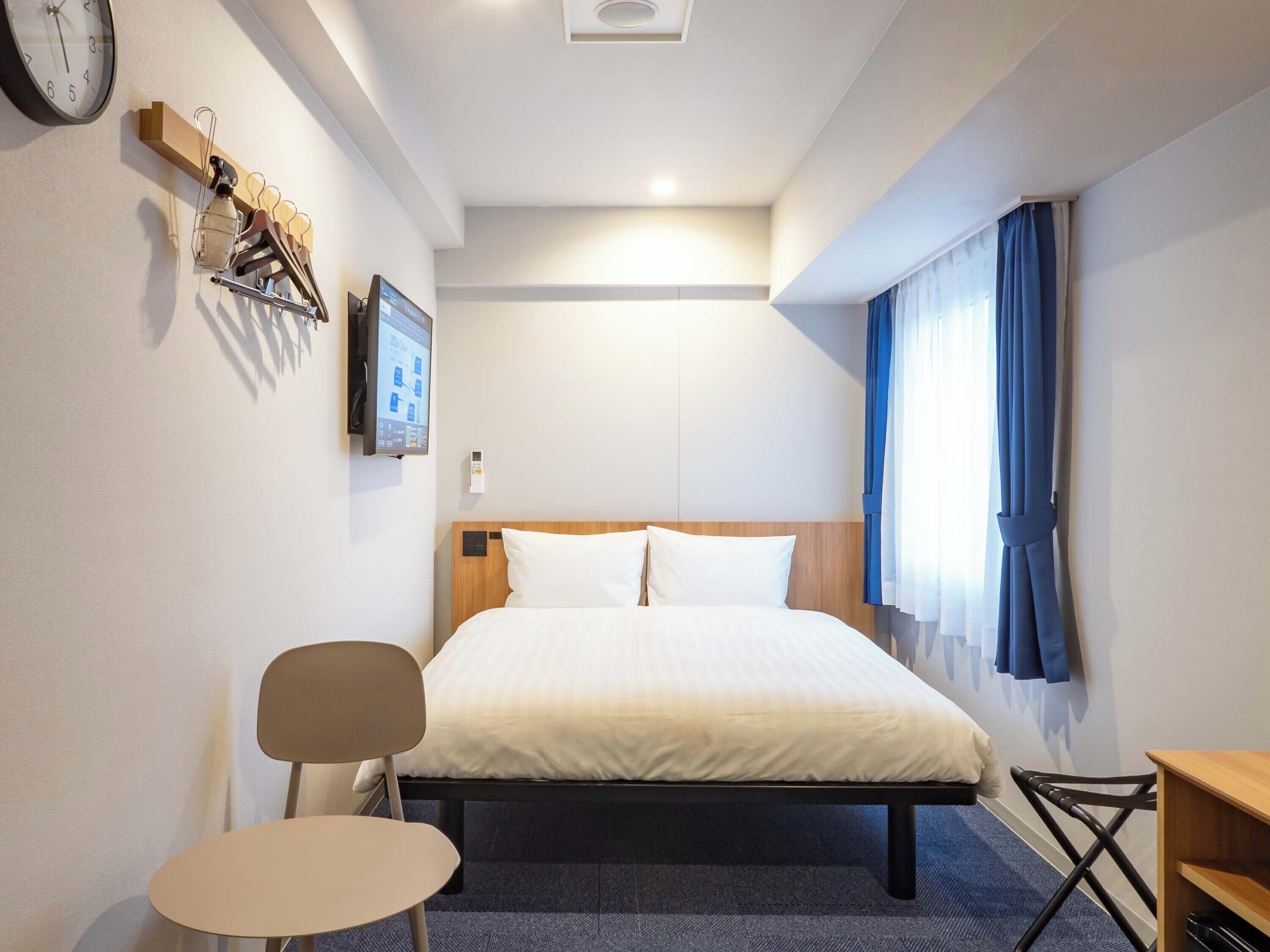 A hotel room with a bed, a desk, a chair, a clock, and a wall-mounted TV.