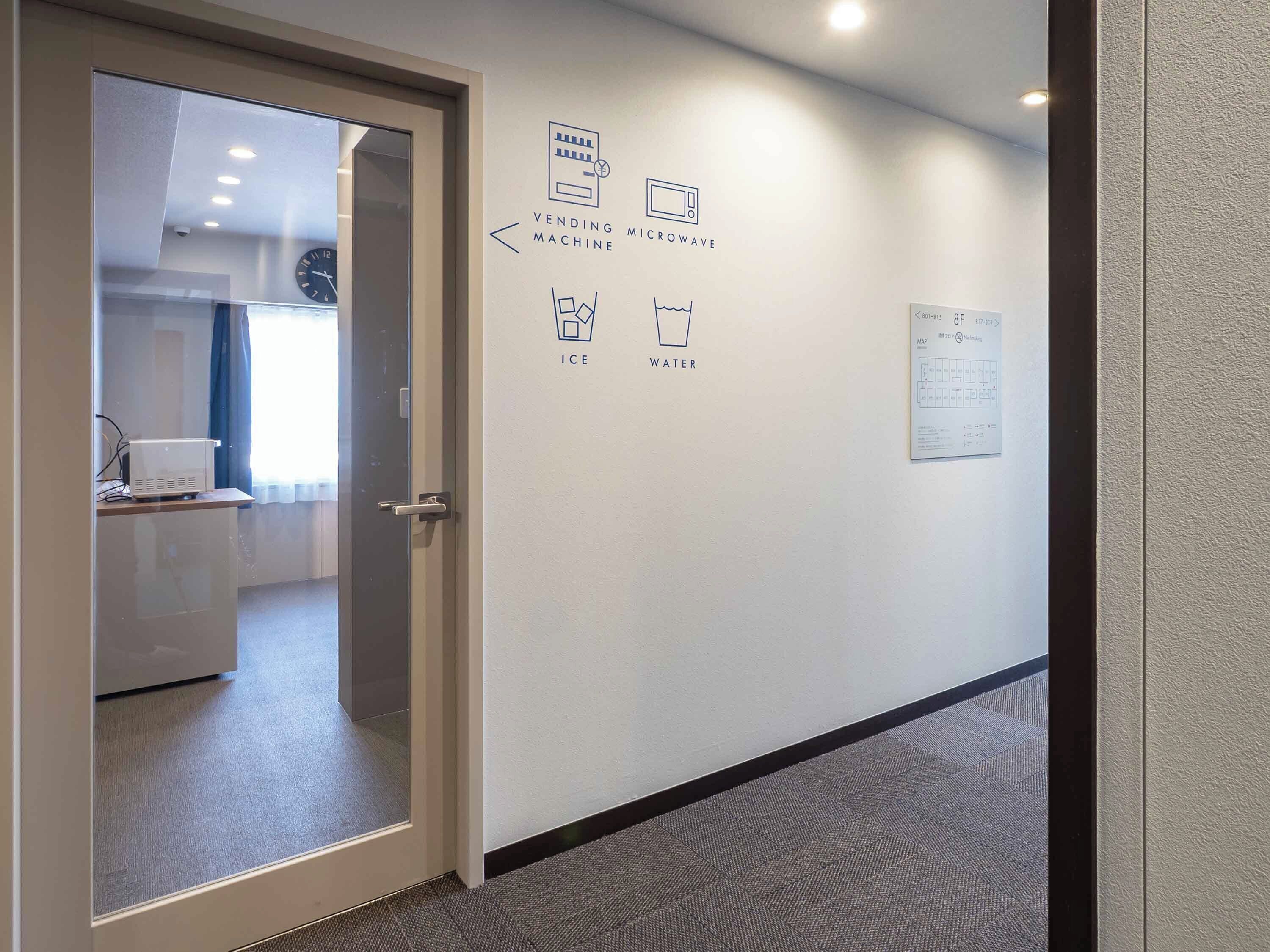 A hallway with a door leading to a room, a wall with icons indicating amenities like a vending microwave, ice, and water, and a map on the wall.