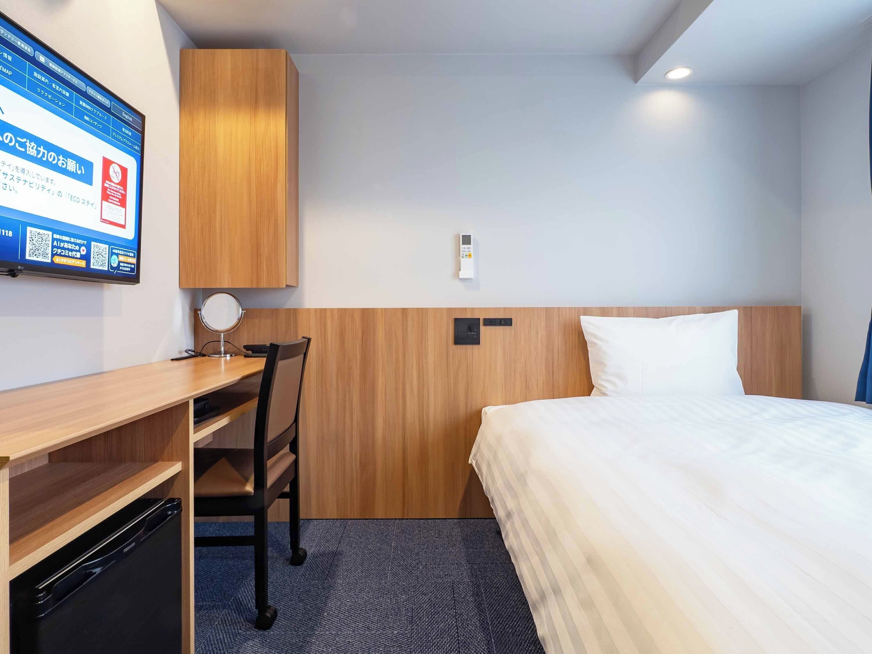 A hotel room with a bed, a desk, a chair, a television, and a wall-mounted control panel.