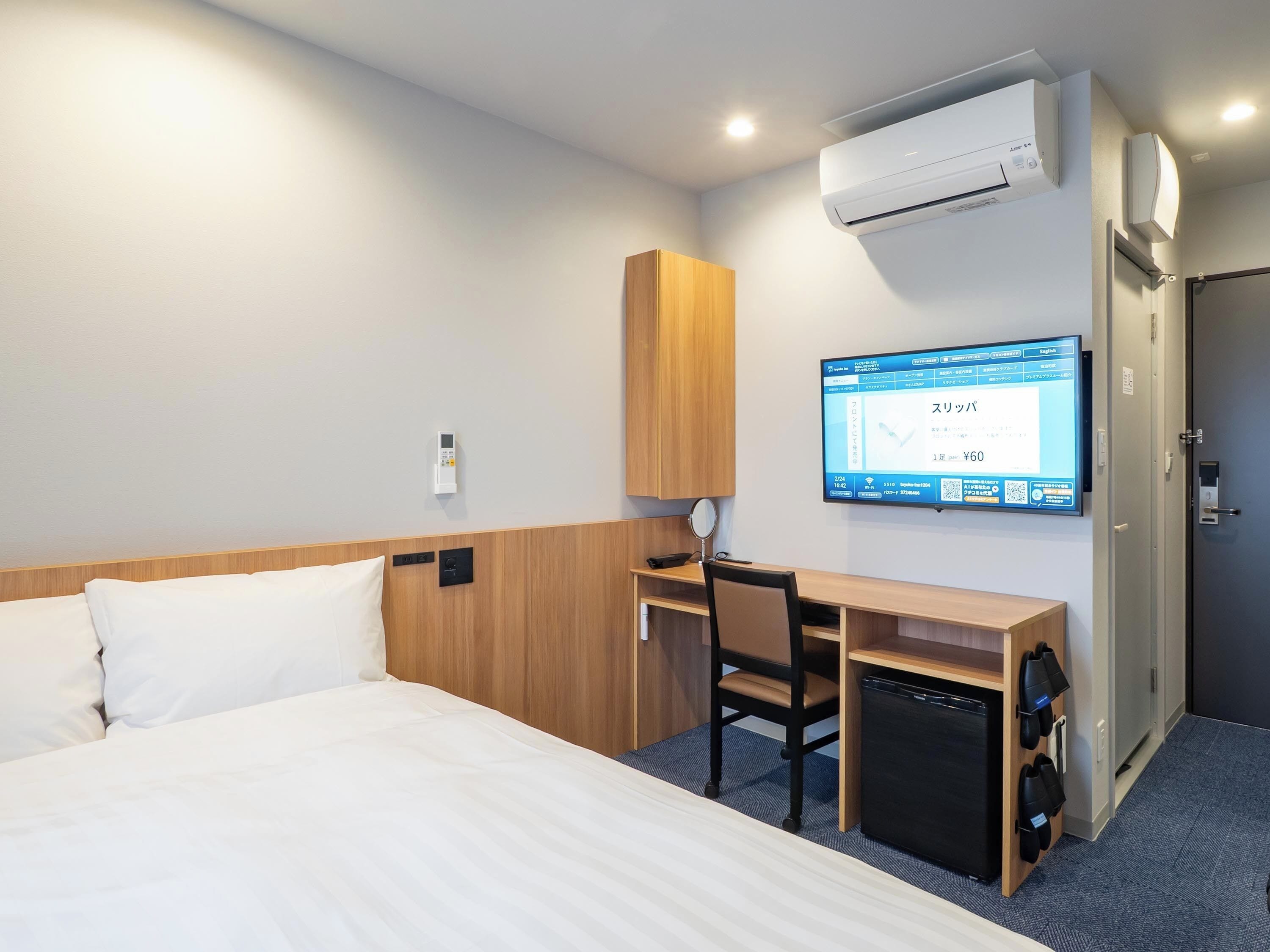 A hotel room with a bed, desk, chair, air conditioning unit, TV, and a door.
