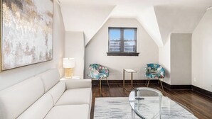 Camera da letto moderna in stile loft con una grande finestra, un divano bianco, due poltrone floreali, un tavolino da caffè in vetro e un'ampia pittura astratta.