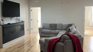 A spacious living room with a grey sofa, a flat-screen TV mounted on the wall, and a ceiling fan.