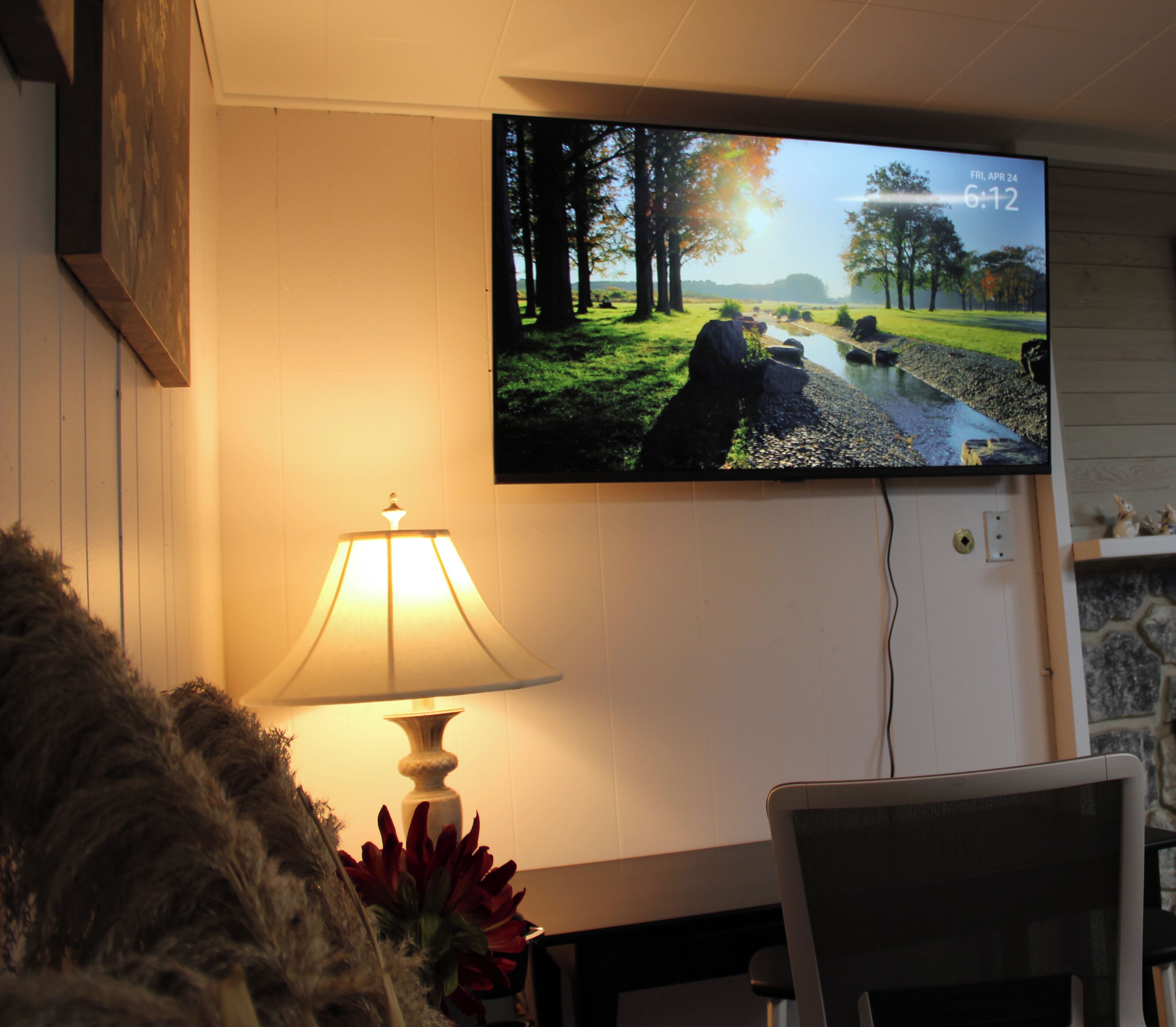 A television mounted on a wall displaying a scenic landscape, a lamp on a table, and a chair.