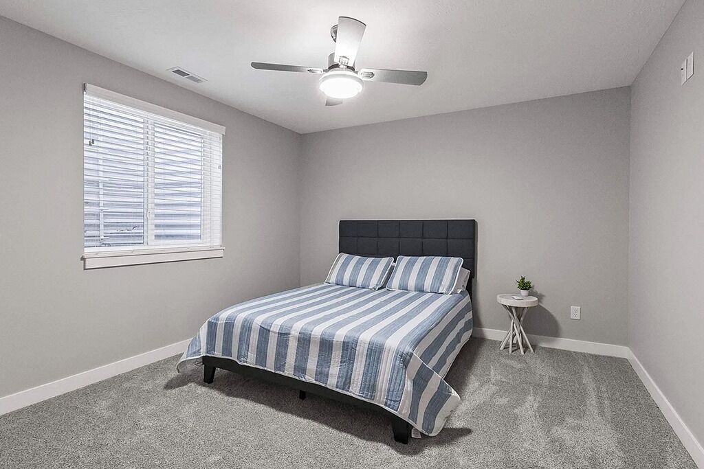 Un dormitorio con cama, ventilador de techo, ventana con persianas y una mesita redonda con una planta.