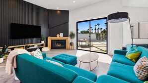 Ein modernes Wohnzimmer mit türkisfarbenen Sofas, einem Flachbildfernseher, einem Kamin und einer großen Schiebeglastür zu einer Terrasse.