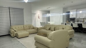 A modern living room with a sectional sofa, a dining area with bar stools, and a kitchen with white cabinets. - Delightful 4-bedroom 4 full bath and one Den house with AC and heater Lehigh  (Lehigh Acres)