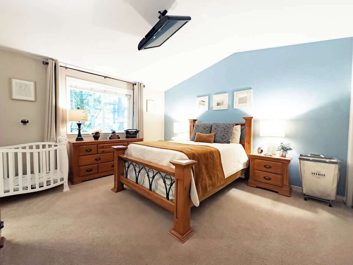 A bedroom with a wooden bed, a nightstand, a dresser, a crib, a window with curtains, and a ceiling light.