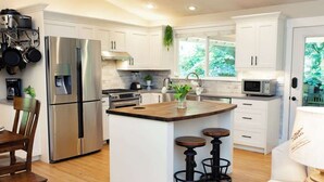 Et moderne køkken med hvide skabe, rustfrie stålapparater, en træø med barstole og udsigt til grønne områder.