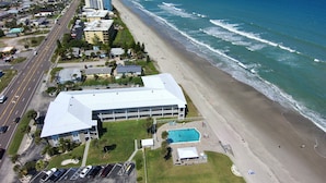 An aerial view of a coastal hotel with a swimming pool, beach access, and a road running parallel to the beach. - Monthly Stay Oceanside Condo — Heated Pool, on the Beach & Fire Pit! 🌴🌊 (Satellite Beach)