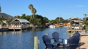 Un muelle frente al agua con sillas Adirondack y una pequeña mesa, rodeado de construcciones de madera y palmeras.