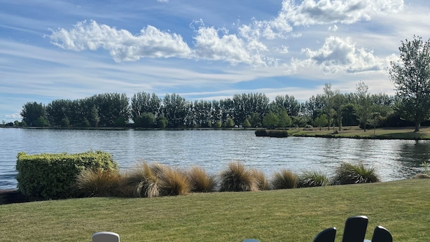 Two Adirondack chairs facing a lake with a grassy area and trees in the background.