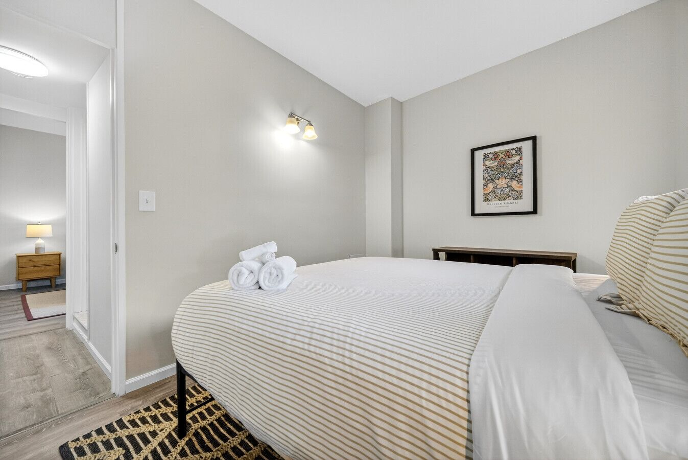 Un dormitorio con cama, mesita de noche, lámpara, un cuadro en la pared y una puerta que da a otra habitación.