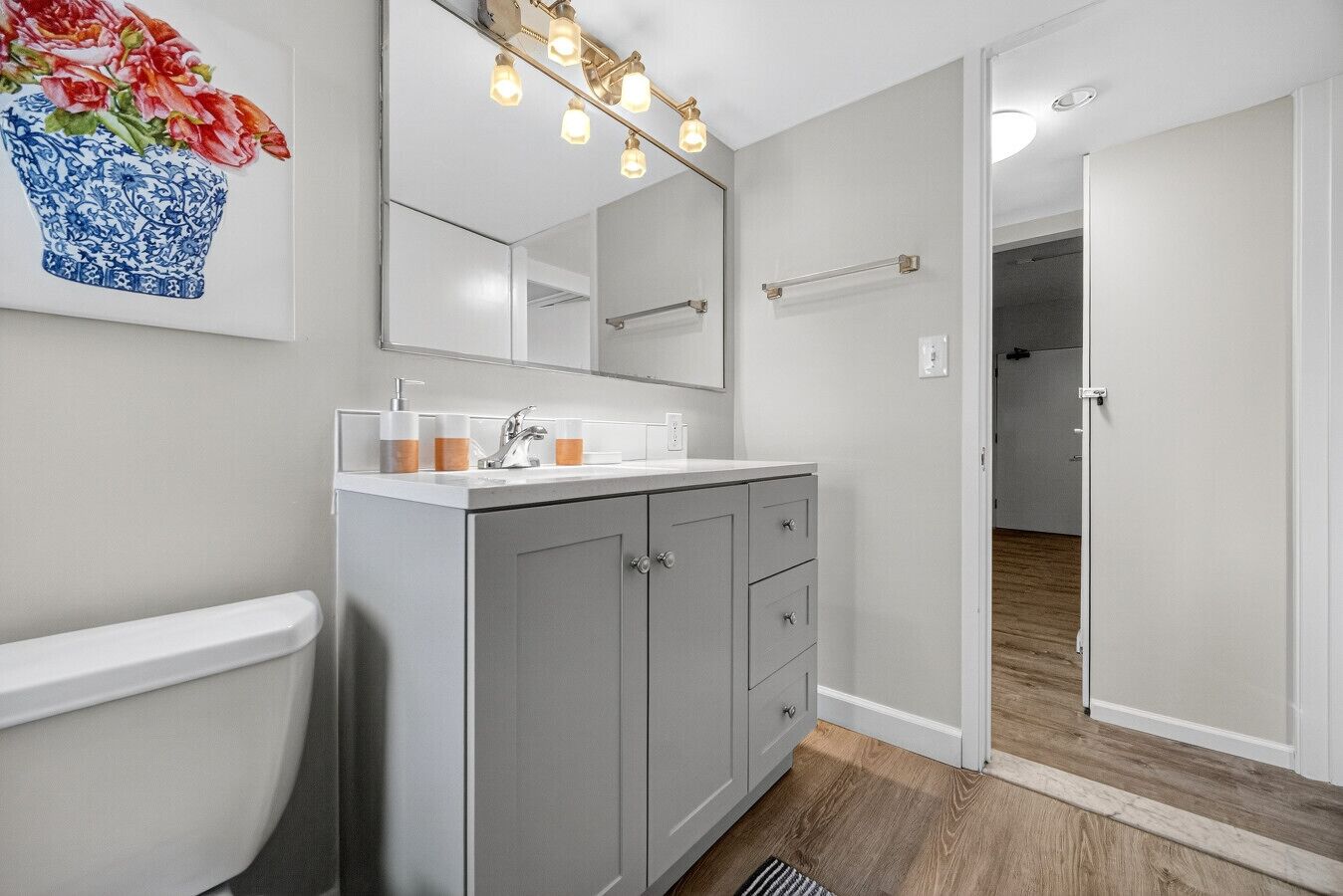 Een moderne badkamer met toilet, wastafel en spiegel. Er ligt een houten vloer en een gang die naar een andere kamer leidt.