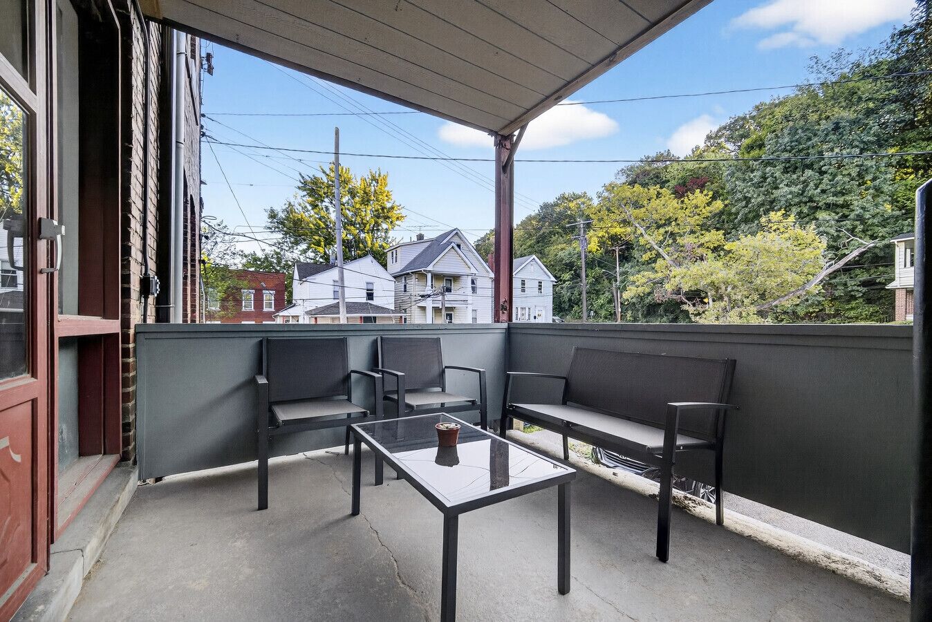 A balcony with four black chairs and a glass table, overlooking a residential area with houses and trees.
