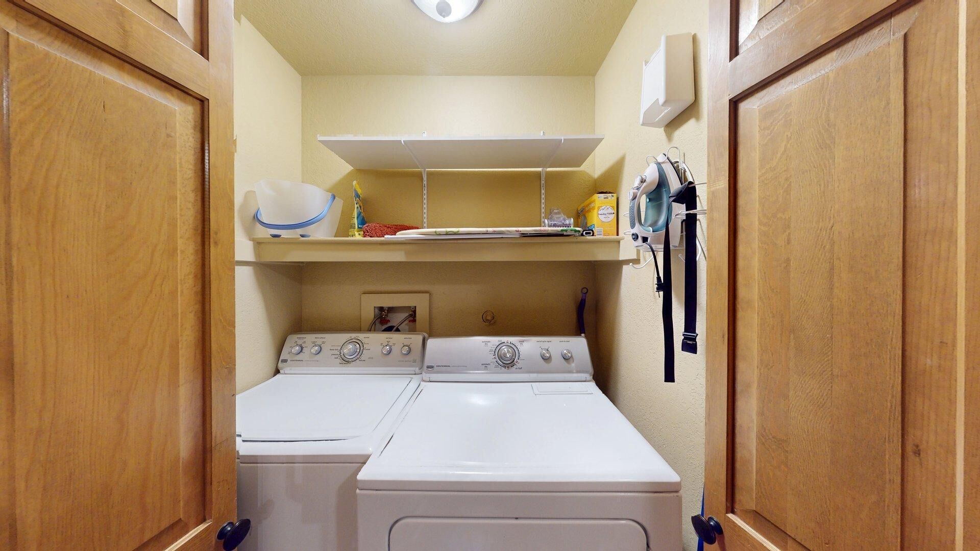 A compact laundry room with two stacked washers and dryers, a shelf above for storage, and a wall-mounted drying rack.