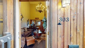 A wooden door with the number 303, leading into a living room with a sofa, a coffee table, and a chandelier. - Walk to Slopes | Master ensuite | Snowfire 303B (Angel Fire)