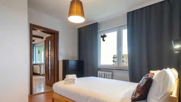 Ein modernes Schlafzimmer mit einem hölzernen Bettgestell, einem großen Fenster mit Vorhängen, einem Heizkörper und einem Fernseher auf einem Schrank.