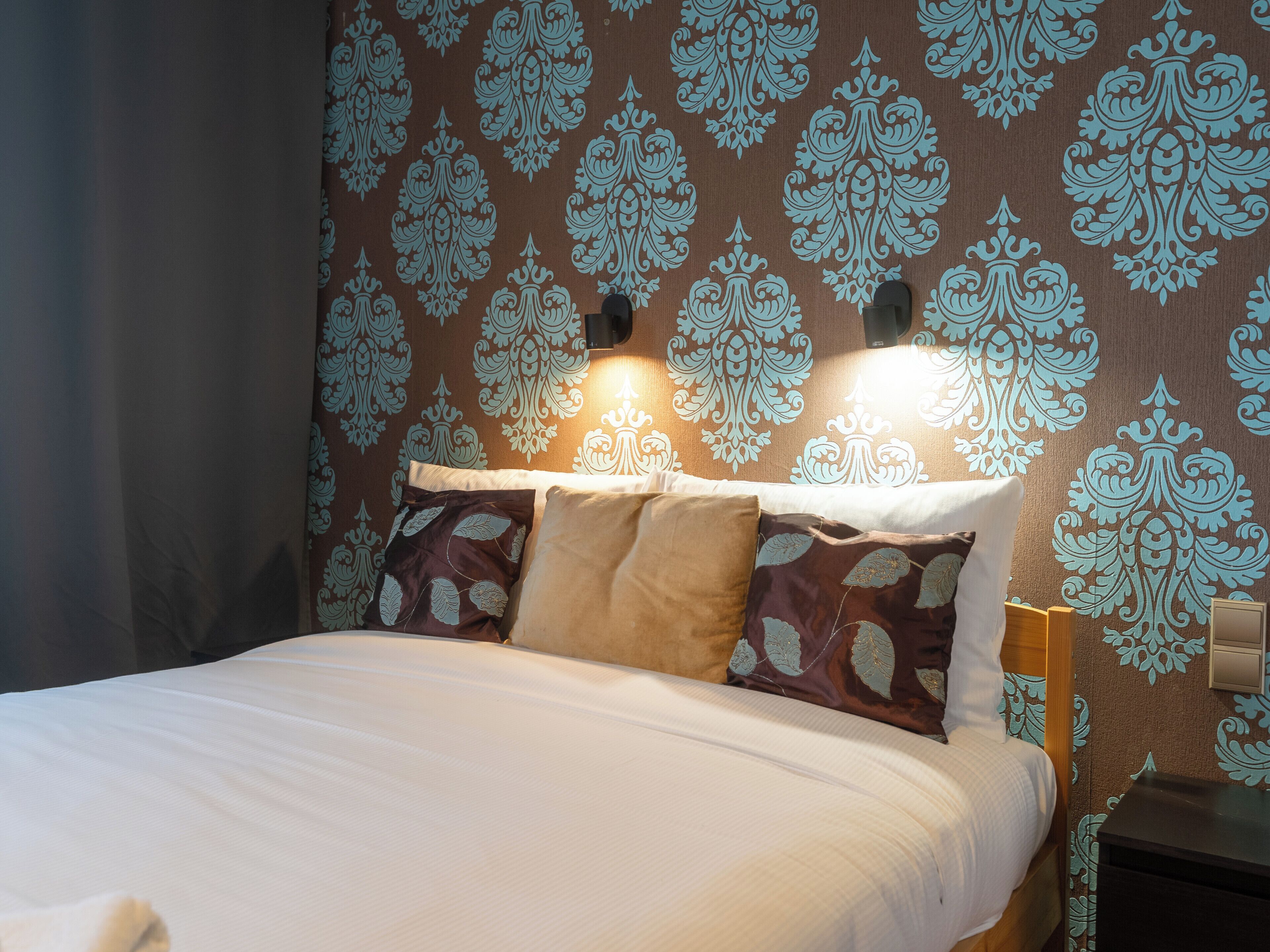 A neatly made bed with patterned pillows, a bedside table, and a wall with a decorative damask pattern.
