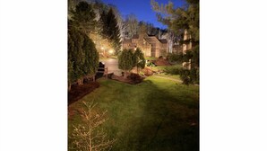 A residential property at dusk with a well-lit driveway, a wooden fence, and a lawn.
