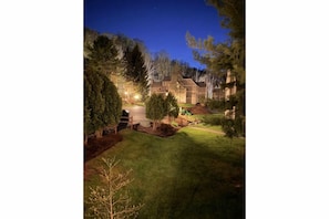 A residential property at dusk with a well-lit driveway, a wooden fence, and a lawn.