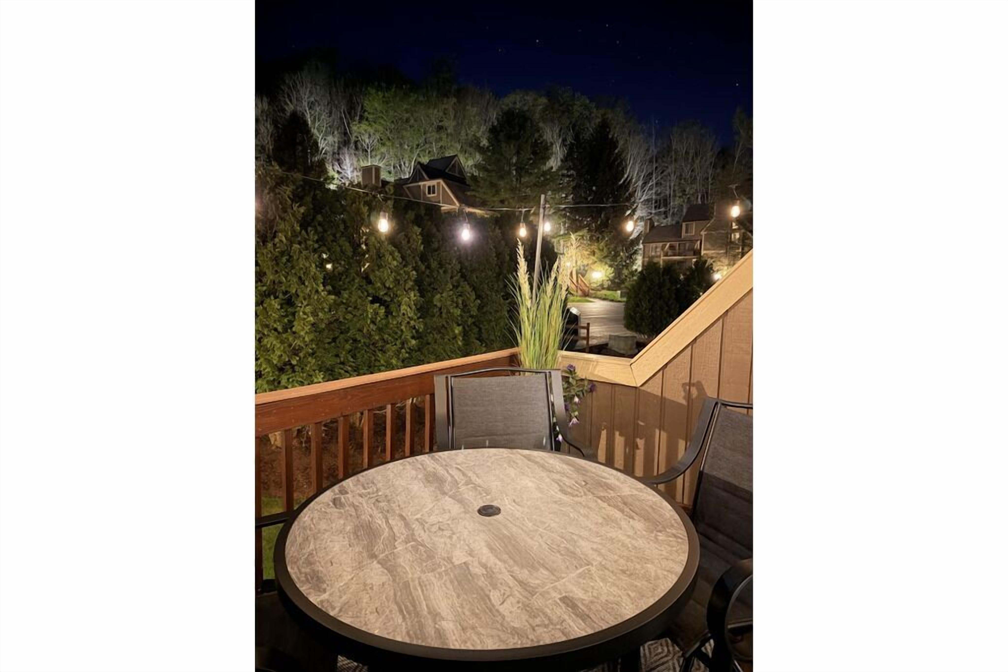 Une table ronde entourée de chaises, placée sur un balcon délimité par une balustrade en bois, avec des guirlandes lumineuses et de la verdure en arrière-plan.