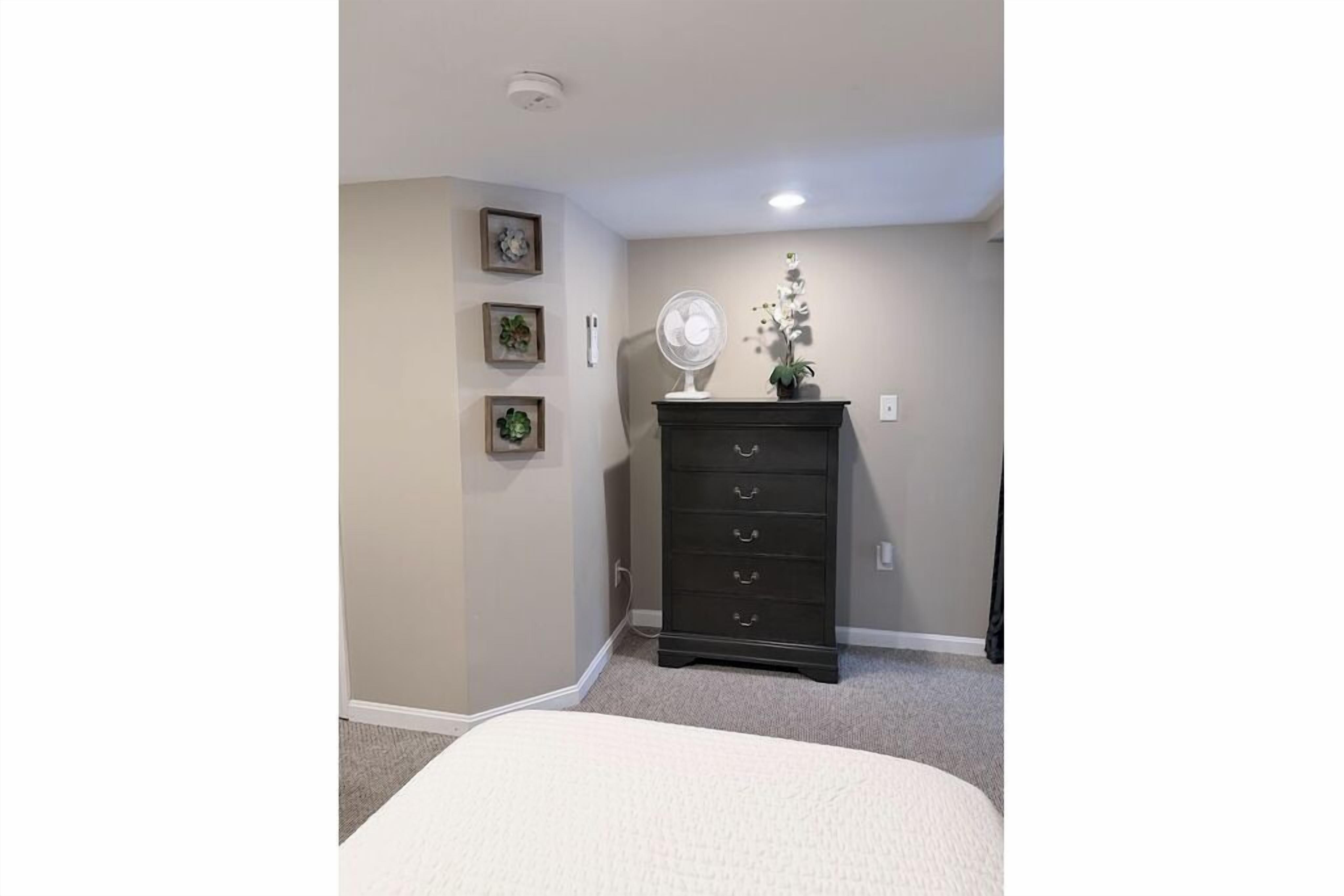 A bedroom with a bed, a dresser, a wall-mounted fan, and framed pictures.