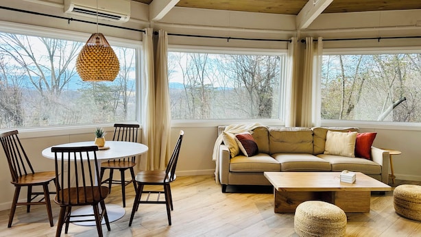 Un vaste espace de vie doté d’un plafond en bois, de grandes fenêtres, d’une table de salle à manger ronde avec des chaises, d’un canapé beige et d’une table basse en bois.