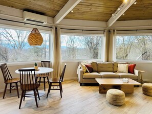 Ein großzügiger Wohnbereich mit Holzboden, großen Fenstern, einem runden Esstisch mit Stühlen, einem beigefarbenen Sofa und einem Couchtisch aus Holz.