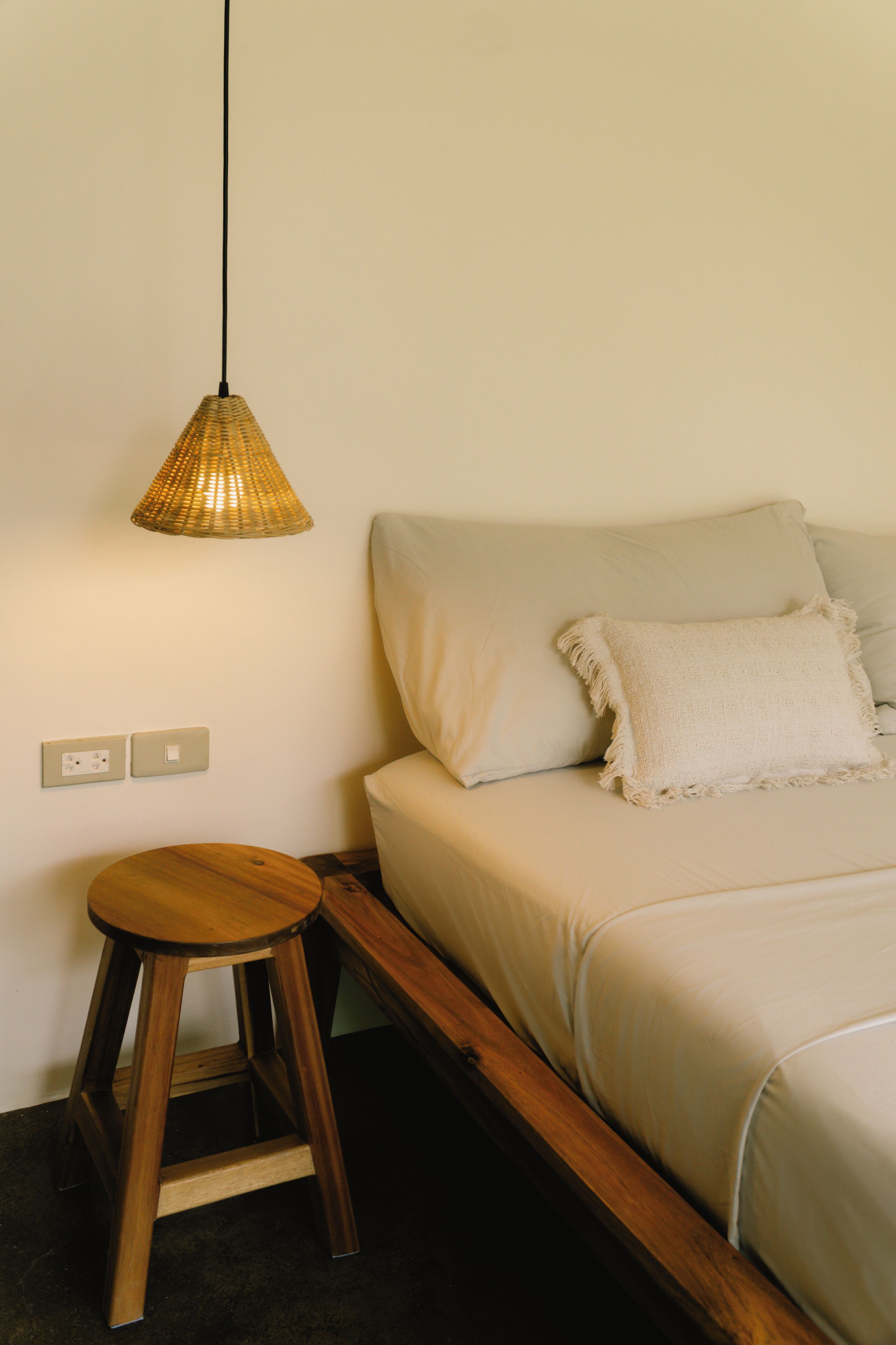 Un dormitorio con una cama, un taburete de madera, una lámpara colgante y un enchufe de pared.