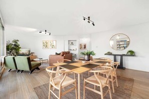 Ein modernes Wohnzimmer mit einem Holzesstisch, Stühlen, einer Couch und einem Couchtisch. Es gibt Pflanzen, Kunstwerke an den Wänden und einen runden Spiegel auf einer Konsolentisch.