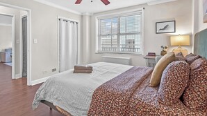 Una camera da letto con un letto, una ventola a soffitto, una finestra con persiane, un comodino con una lampada e un quadro appeso al muro.