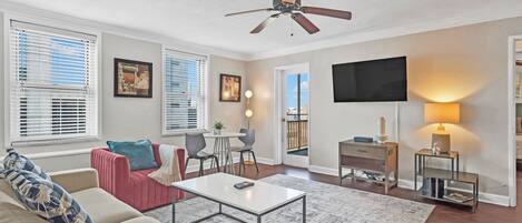 A modern living room with a ceiling fan, a TV mounted on the wall, a dining area with a table and chairs, and a view of the outdoors through the windows.