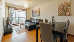 Una sala moderna con un sofá seccional, una mesa de comedor con sillas, una pequeña mesa de centro, un ventanal con cortinas y un cuadro en la pared.