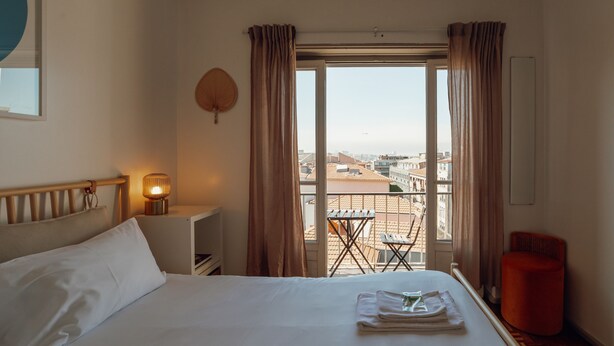 A hotel room with a bed, a bedside lamp, a window with curtains, a small table, and a chair. - Santa Catarina Flat 2BD With Balcony by Vacationy (Porto)