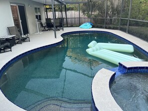 Ein Schwimmbecken mit gefliestem Rand, eine überdachte Terrasse mit Sitzgelegenheiten und ein Holzzaun im Hintergrund.