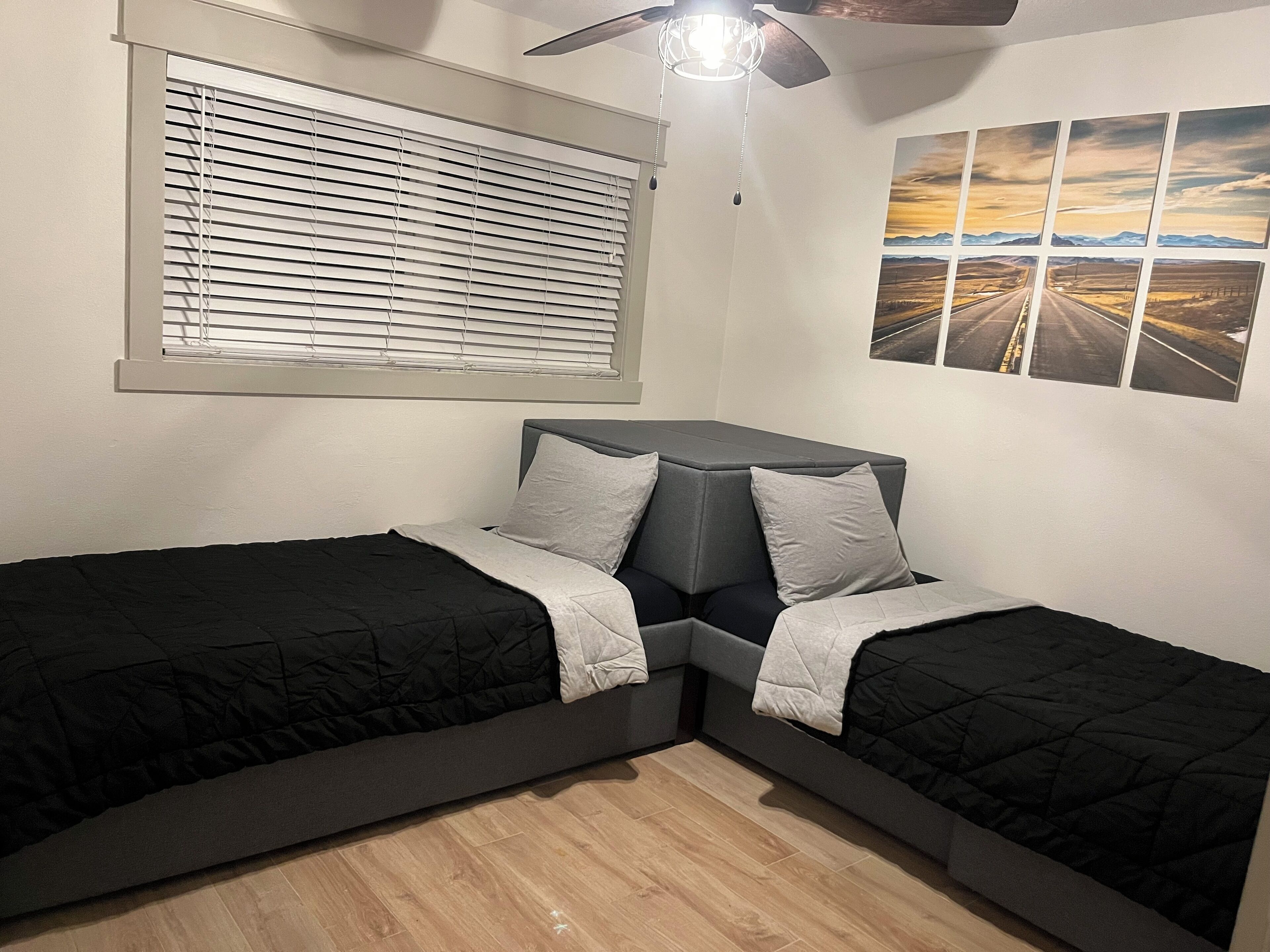Camera da letto con due letti, una ventola a soffitto, una finestra con persiane e un quadro appeso al muro.