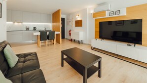 A modern living room with a sofa, a coffee table, and a TV mounted on a wooden wall. There is a kitchen area with white cabinets and a dining table with chairs.