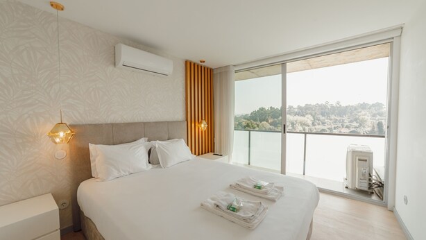 A hotel room with a large bed, a bedside table, a nightstand, a wall-mounted air conditioner, and a view of a body of water through a sliding glass door.