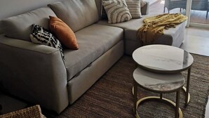 A modern living room with a grey sofa, a round coffee table, and a large window offering a view of outdoor furniture. - Beachfront Studio,  New & Cozy,   Stunning Ocean Views,  Prime Location (Puerto Vallarta)