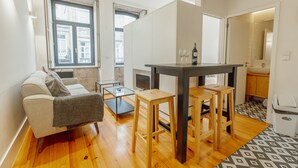 Un espace de vie compact comprenant un canapé, une petite table et un coin cuisine visible à travers une porte ouverte.