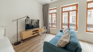 Moderni olohuone, jossa on litteänäyttöinen televisio, lattiavalaisin, sininen sohva ja suuret ikkunat, joista on näkymä rakennuksiin.