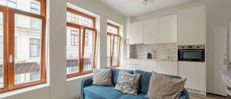 A modern apartment with a blue sofa, white kitchen, and large windows with wooden frames.