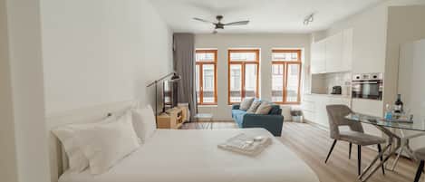 A modern hotel room with a large bed, a small sofa, a glass dining table, and a kitchenette.