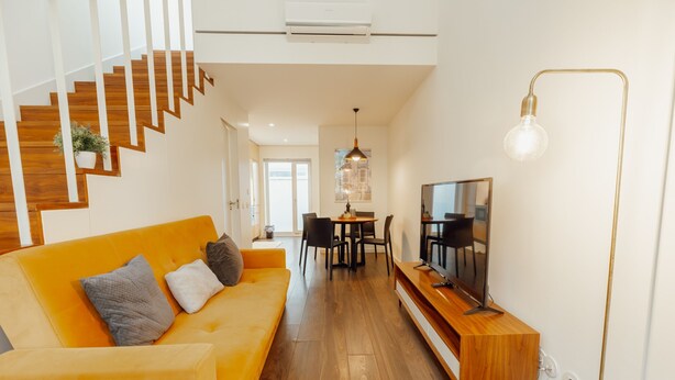 Ein modernes Wohnzimmer mit einer Holztreppe, einem leuchtend gelben Sofa, einem Essbereich und einem Fernseher auf einem Holzboden.