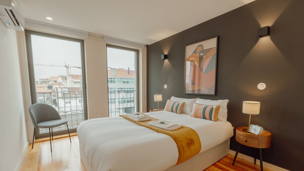 Een moderne hotelkamer met een groot bed, een stoel, een nachtkastje met lamp en uitzicht op de stad door grote ramen.