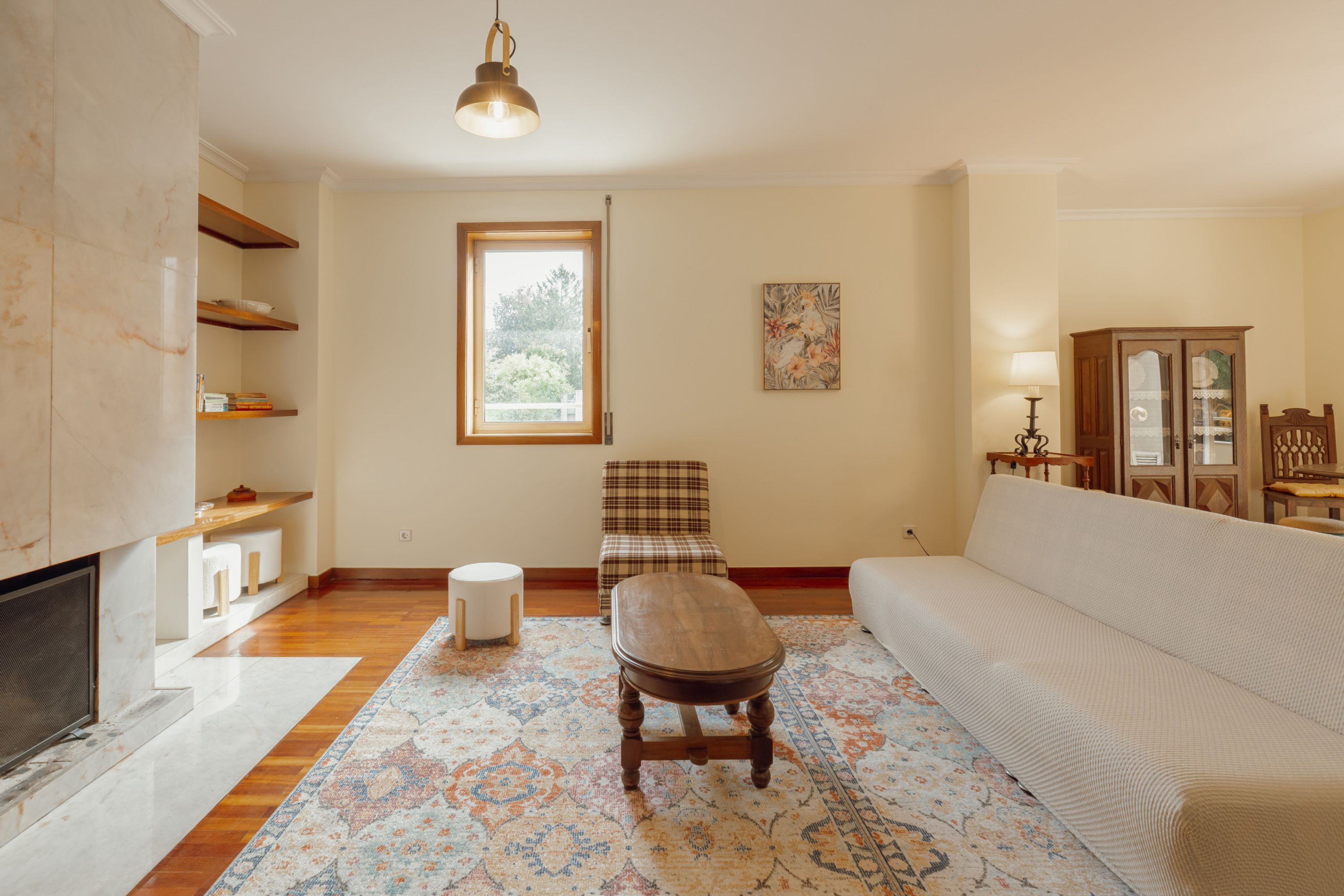 A living room with a sofa, a wooden bench, a patterned rug, a fireplace, and a window with a view of greenery.