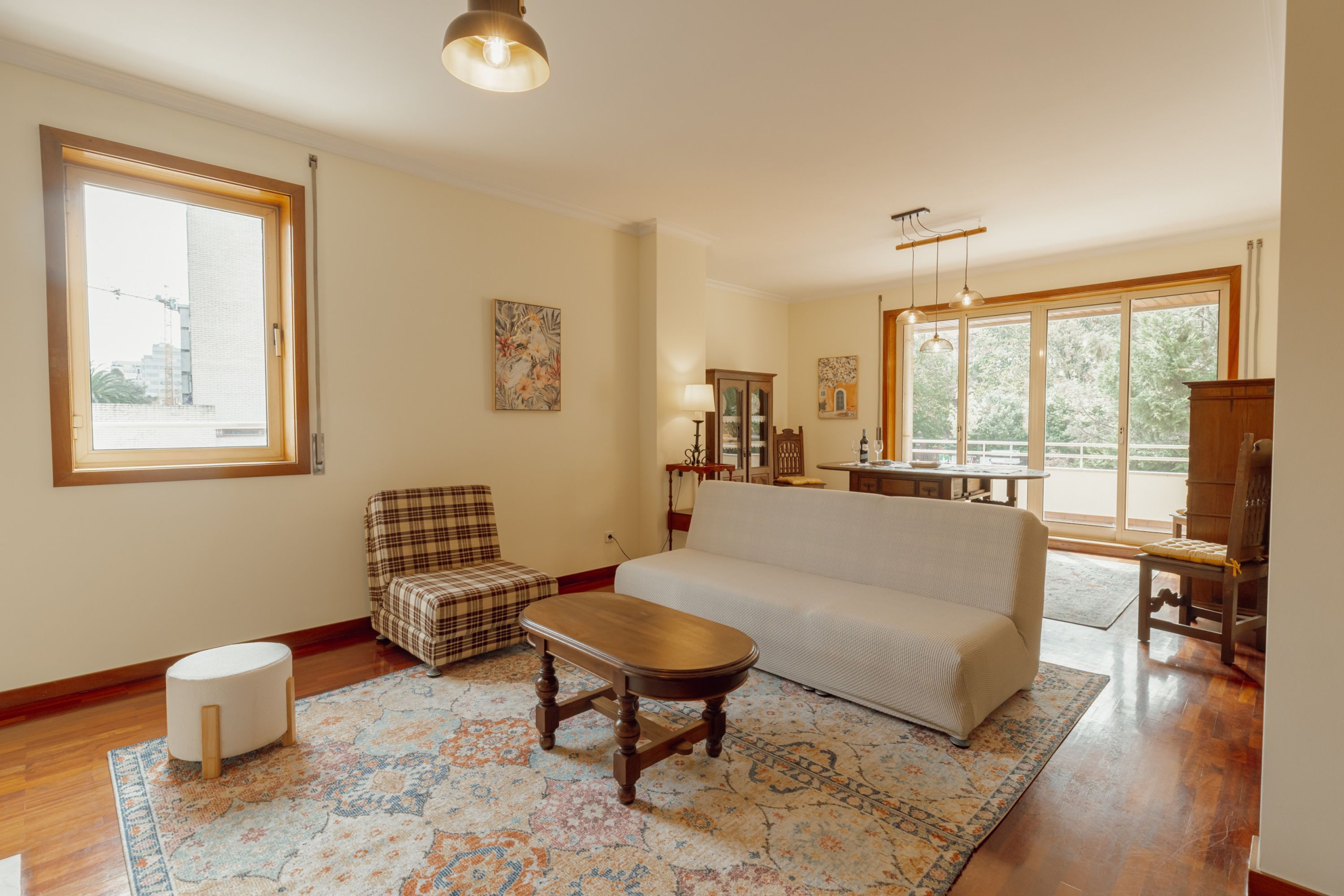 A spacious living room with a large window, a sofa, a wooden coffee table, and a plaid armchair.