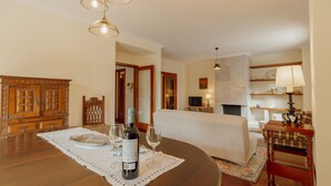 Un comedor con una mesa de madera, una botella de vino y copas. Se observa un sofá y un televisor al fondo.