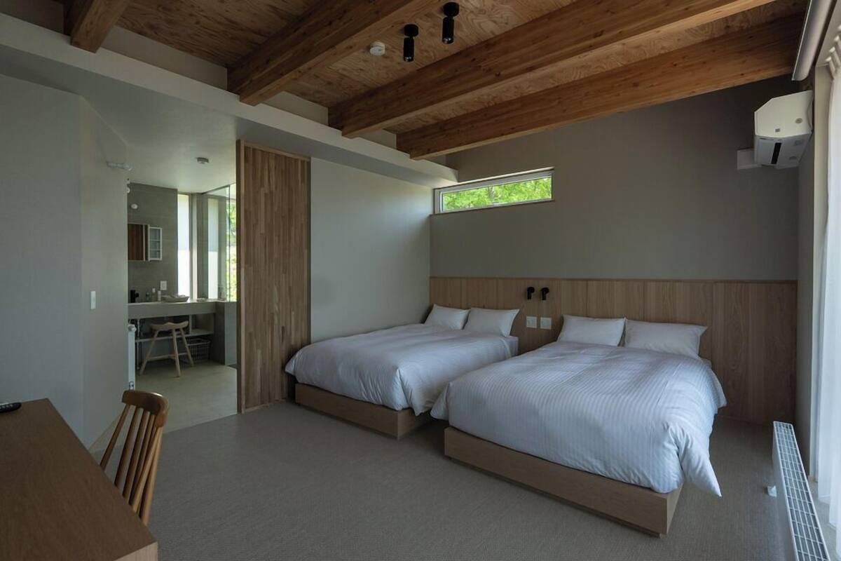 A bedroom with two beds, a wooden ceiling, a window, and a bathroom visible through an open door.
